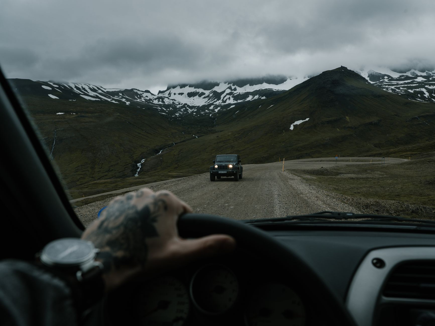 Main tatouée sur le volant, vue depuis l’habitacle, face à une route de gravier en montagne et à un 4x4 qui approche.
