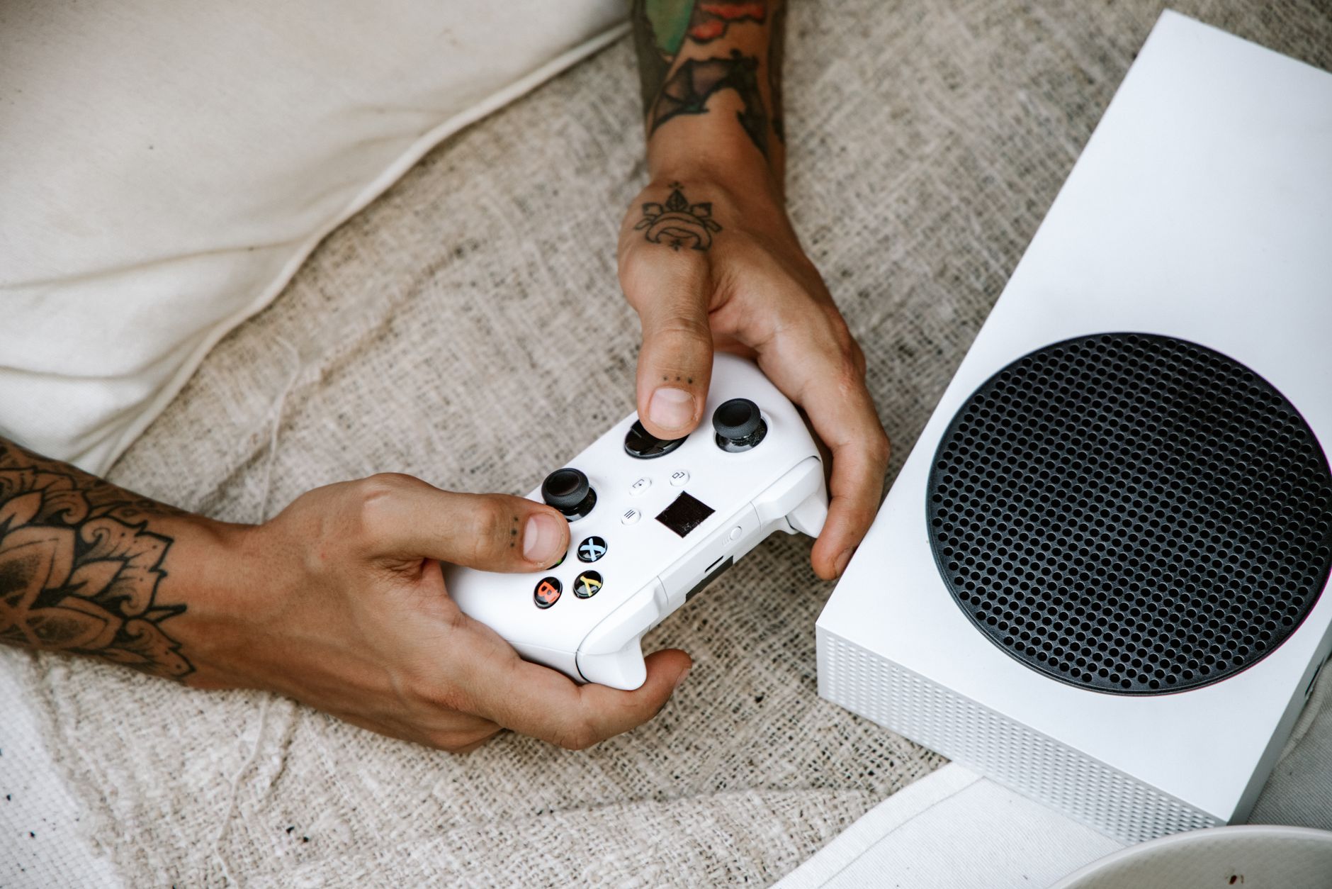 Mains tenant une manette de jeu vidéo blanche à côté d’une console posée sur un drap.