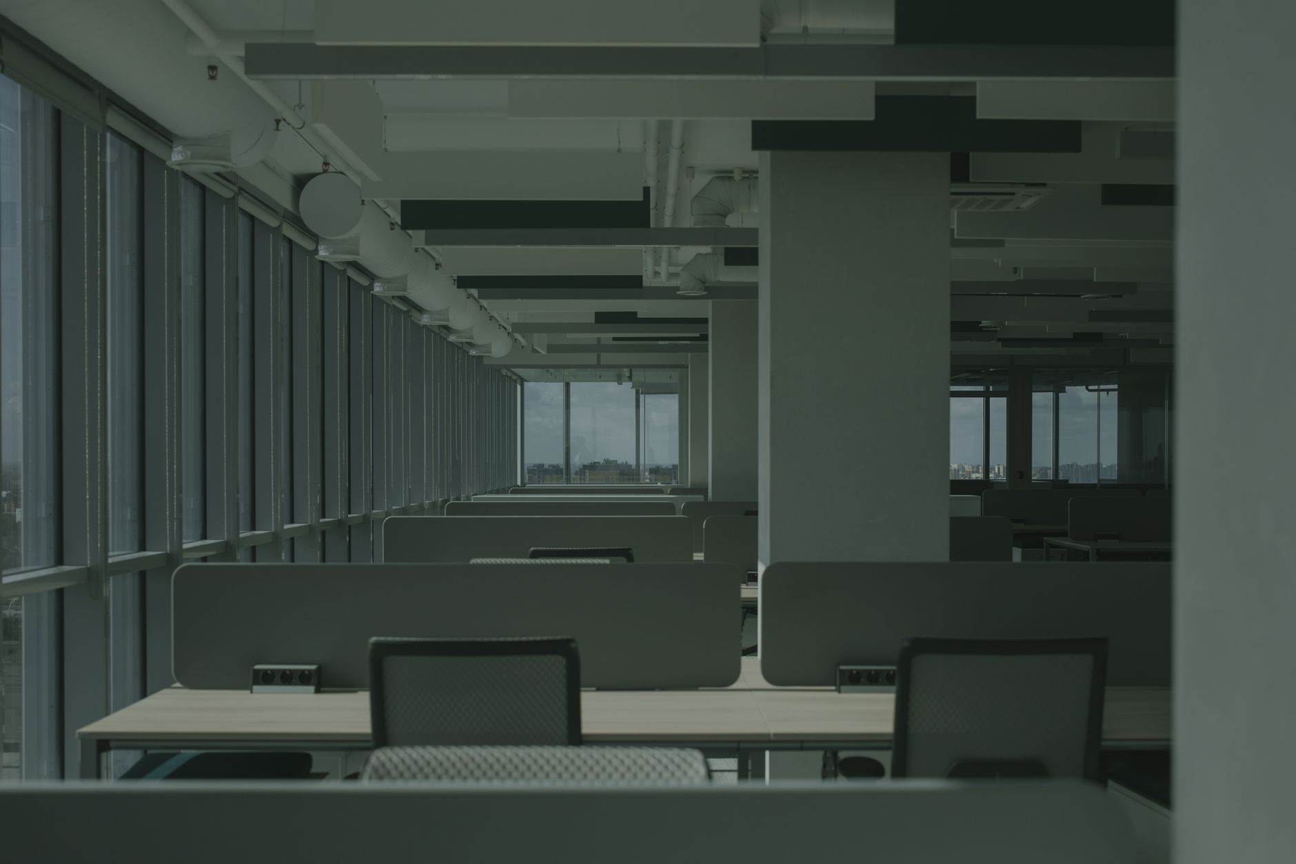 Open space vide en lumière sombre, avec rangées de bureaux, chaises, cloisons basses et grandes baies vitrées donnant sur la ville.