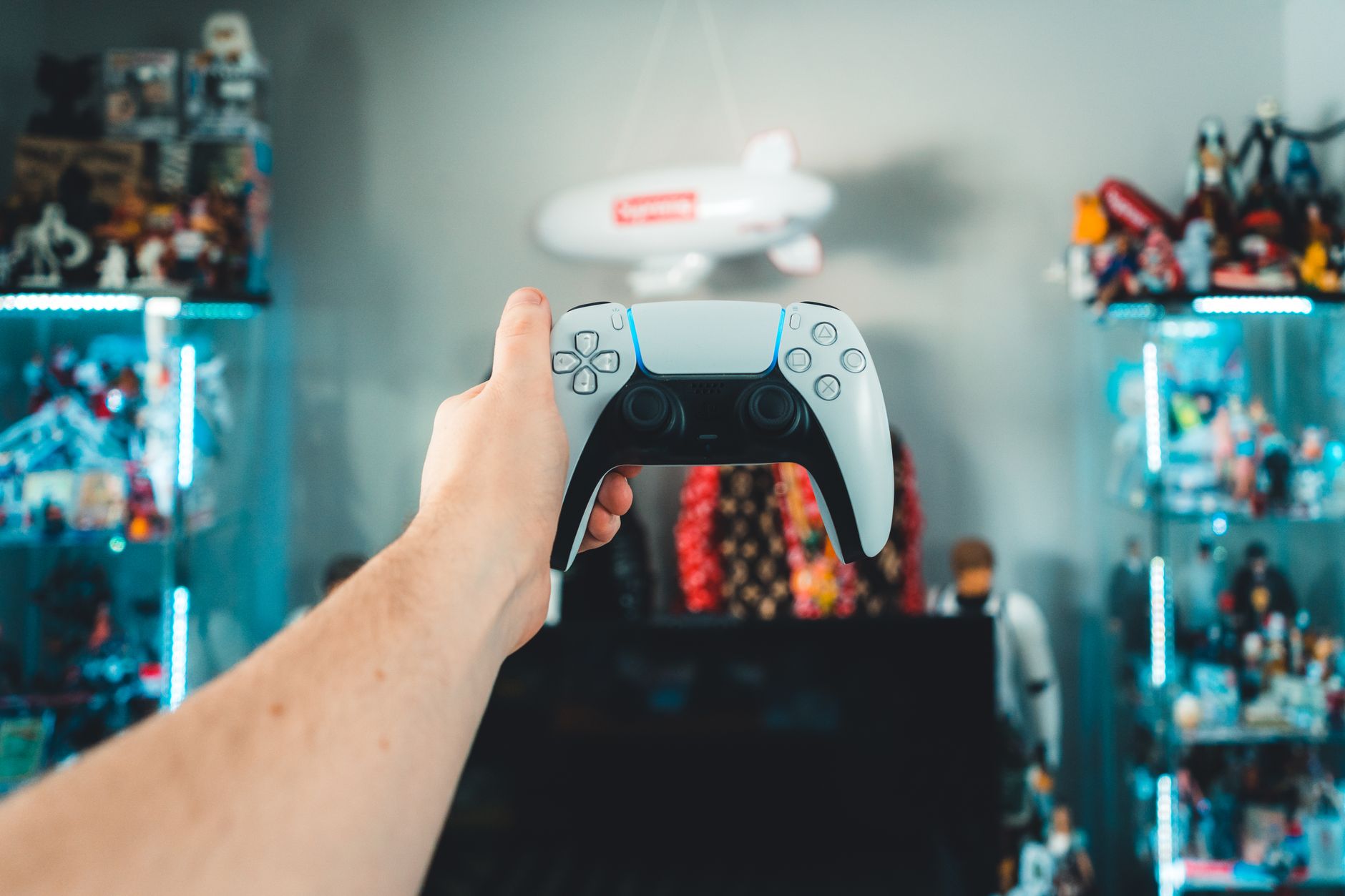 Une main tient une manette de jeu blanche et noire devant un téléviseur, avec des vitrines de figurines éclairées en arrière-plan flou.